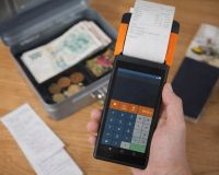 Kädessä pidettävä maksupääte ja avattu kassalipas seteleineen.